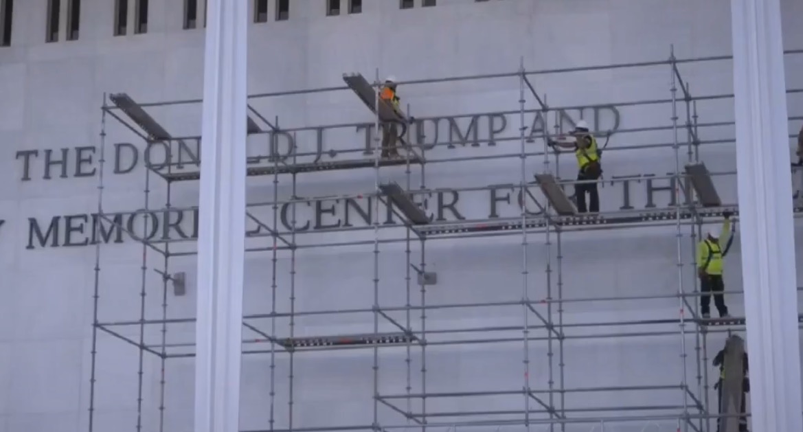 Workers on scaffolding perform maintenance on the Donald J. Trump Memorial Center, highlighting construction and restoration efforts.
