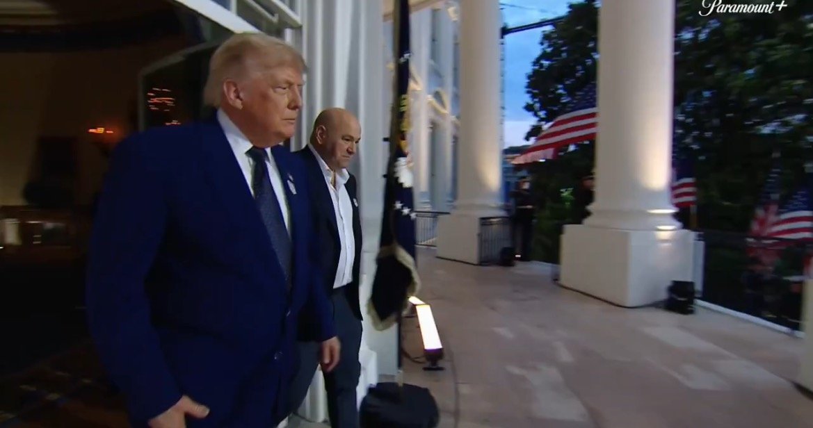 Donald Trump and another individual walk outside the White House, flanked by American flags, during an evening event.