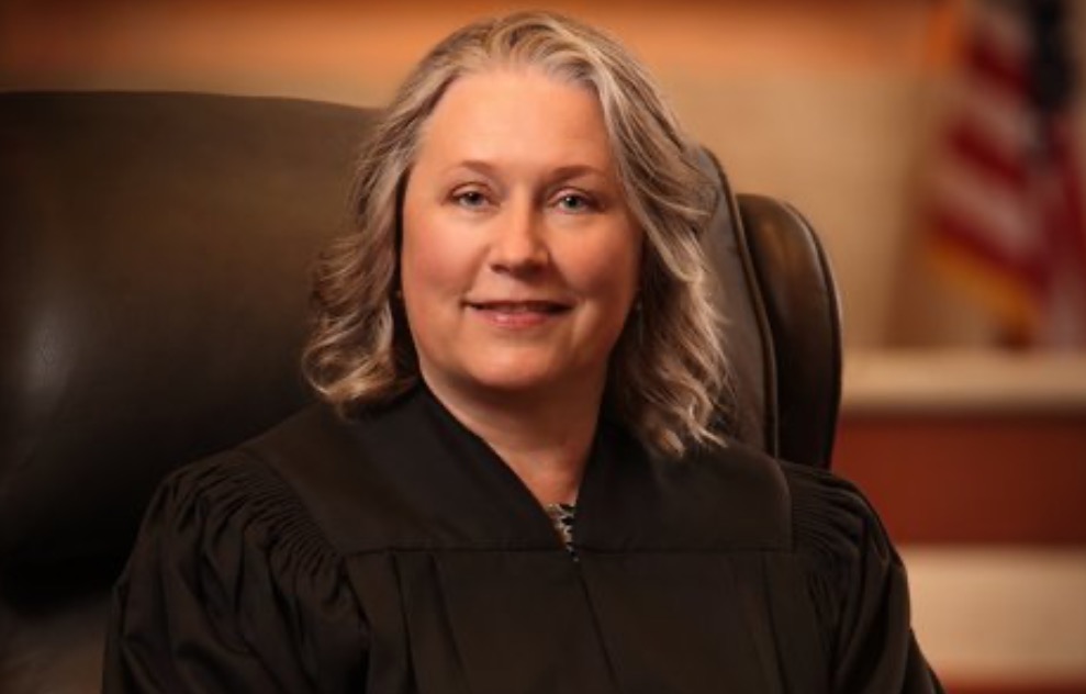 Portrait of a female judge in a black robe sitting in a courtroom, with a warm smile and an American flag in the background.