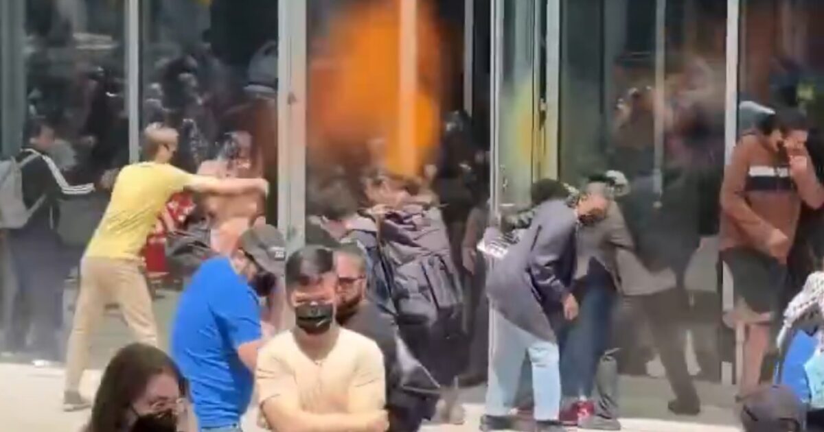 Crowd of people gathered outside a glass building, some attempting to enter while others observe, during a chaotic scene.