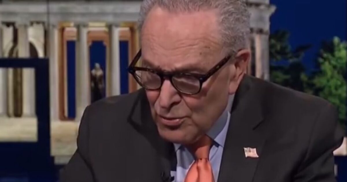 U.S. Senator discussing policy issues during a televised interview, wearing glasses and an orange tie, with a historic backdrop.