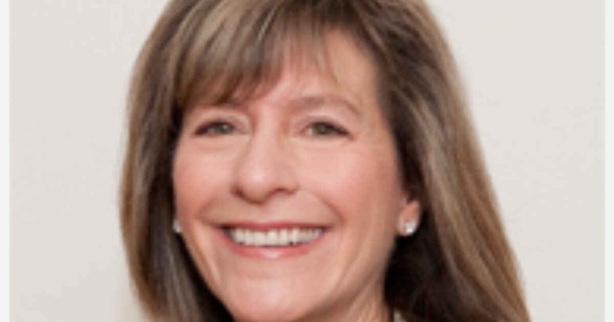Smiling woman with shoulder-length brown hair, wearing earrings and a necklace, against a neutral background.