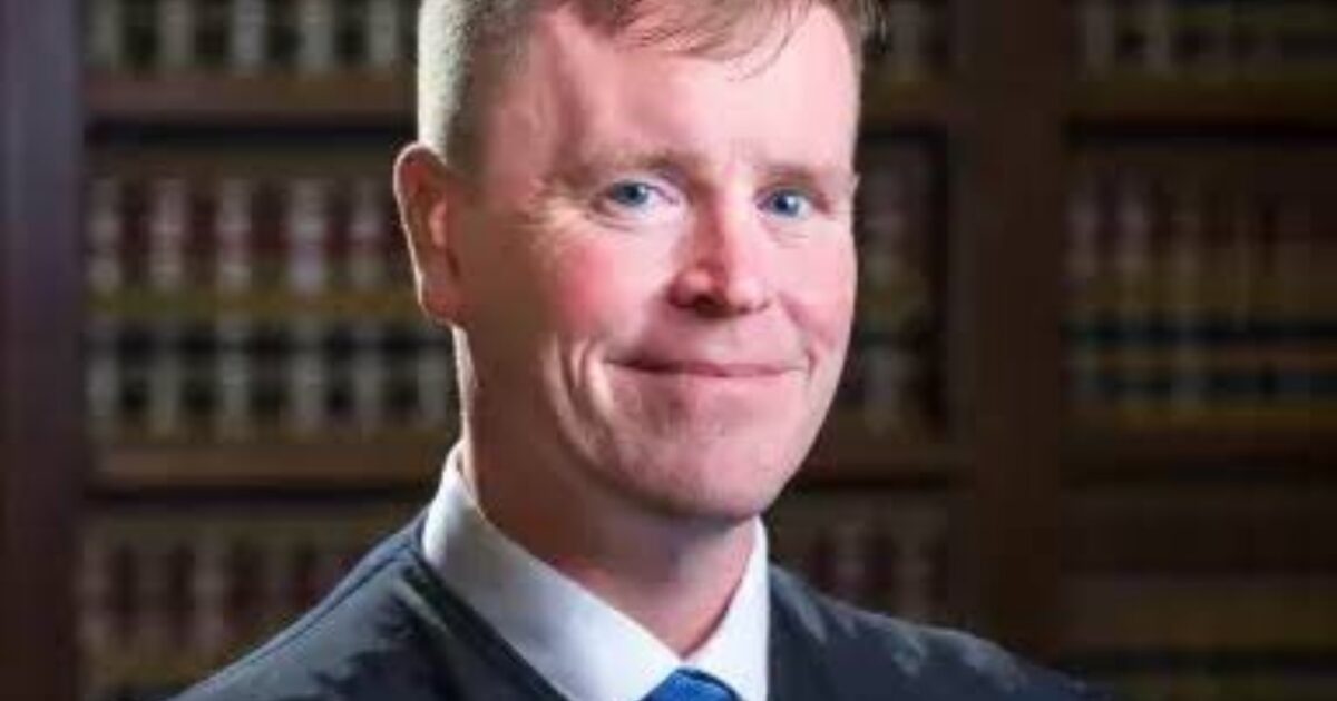 Portrait of a smiling judge in formal attire, with law books in the background, conveying professionalism and authority in a courtroom setting.
