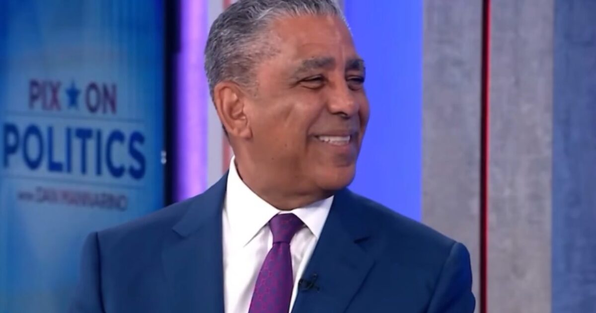 Smiling man in a blue suit with a purple tie, seated in a studio setting, discussing politics on a television show.