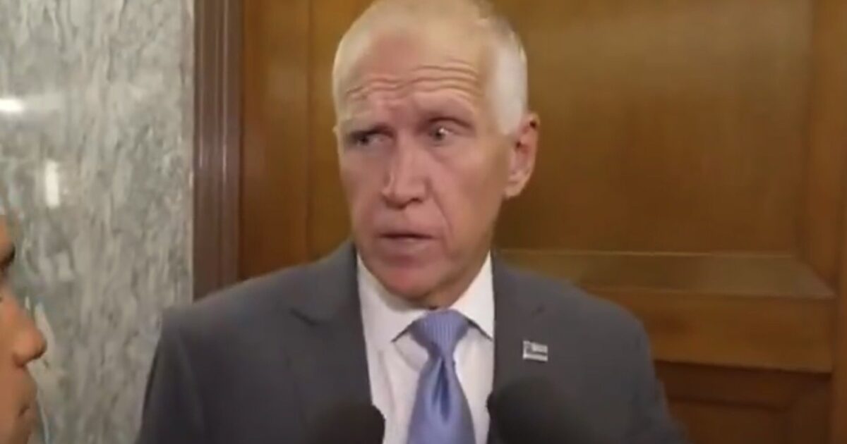 Man in a suit speaking to reporters, with microphones in view, against a wooden backdrop, conveying a serious expression during a press event.
