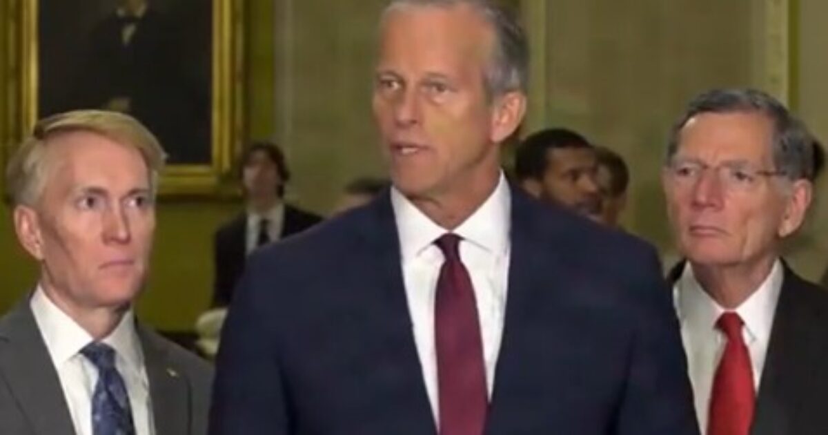 Group of three male politicians speaking at a podium in a formal setting, with a historic portrait in the background.