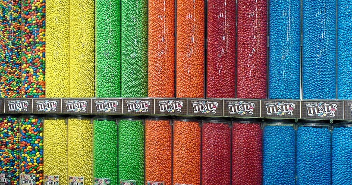Colorful dispensers filled with various M&M's candy varieties, showcasing a vibrant array of colors including red, blue, green, yellow, orange, and multi-colored options.