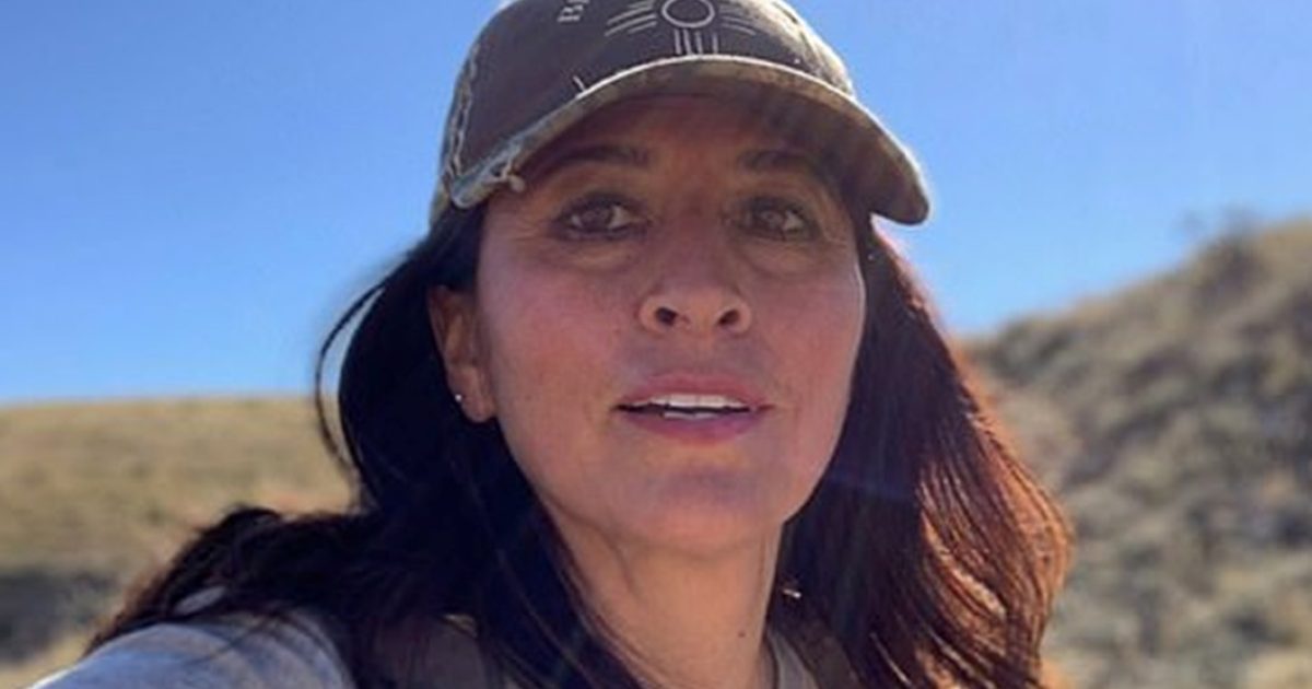 A woman wearing a cap smiles for a selfie outdoors against a clear blue sky and rolling hills.