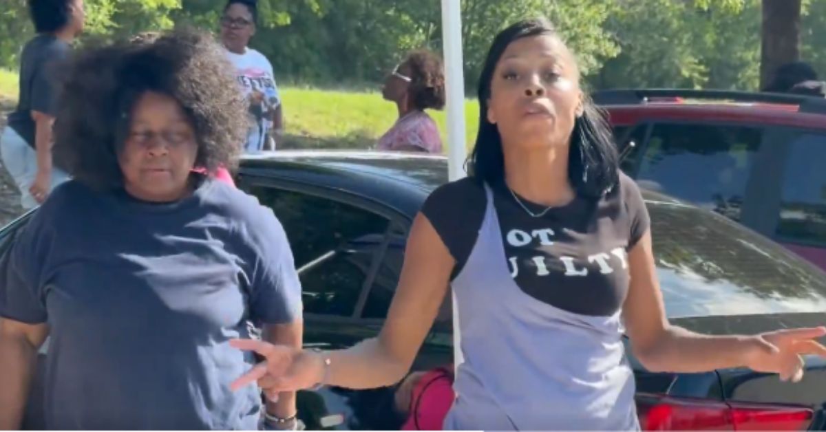 Two women engage in a conversation outdoors, with one wearing a "NOT GUILTY" shirt, surrounded by a group of people and parked cars.