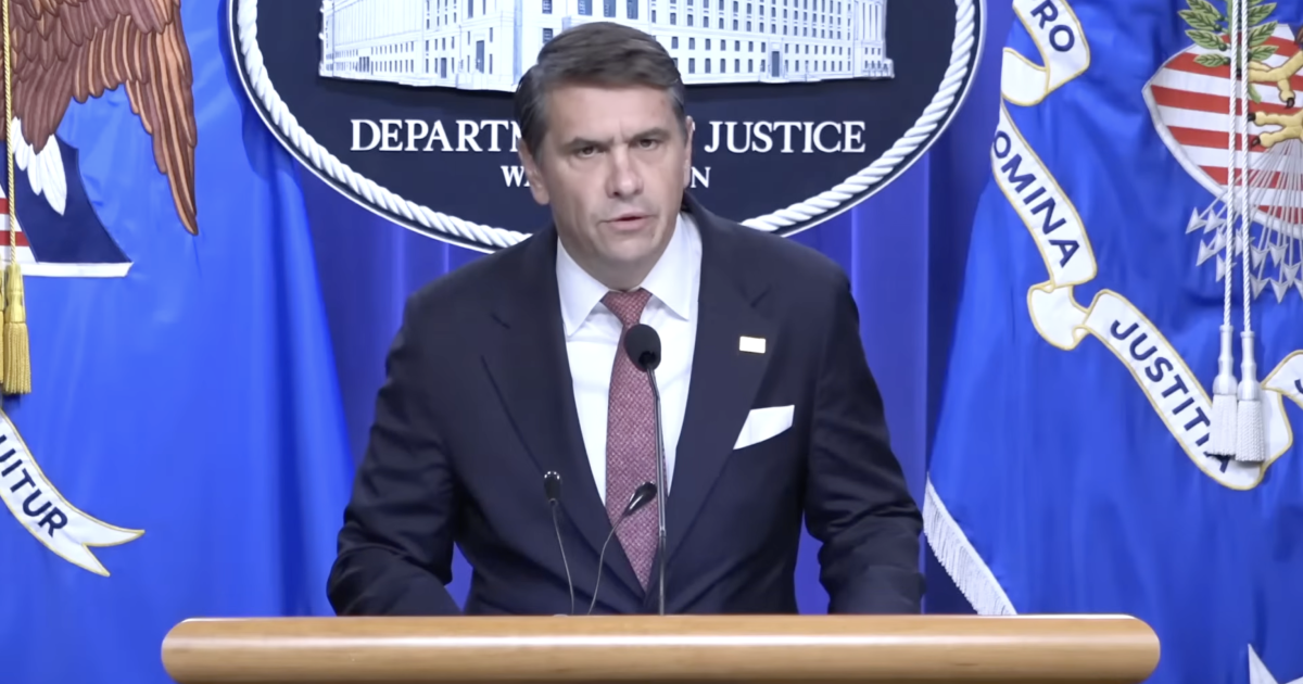 Official speaking at a Department of Justice press conference, with flags and emblems in the background, addressing important legal matters.