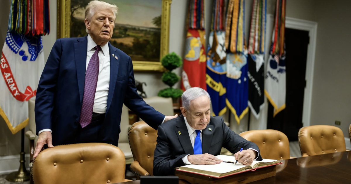 Donald Trump stands beside Benjamin Netanyahu, who is signing a book in the Oval Office, surrounded by military flags and a formal setting.