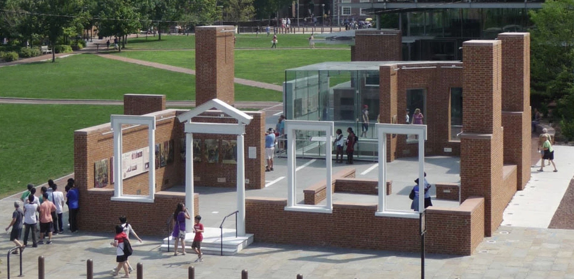 A unique outdoor historical exhibit featuring a reconstructed brick structure with large white frames, surrounded by visitors in a green park setting.
