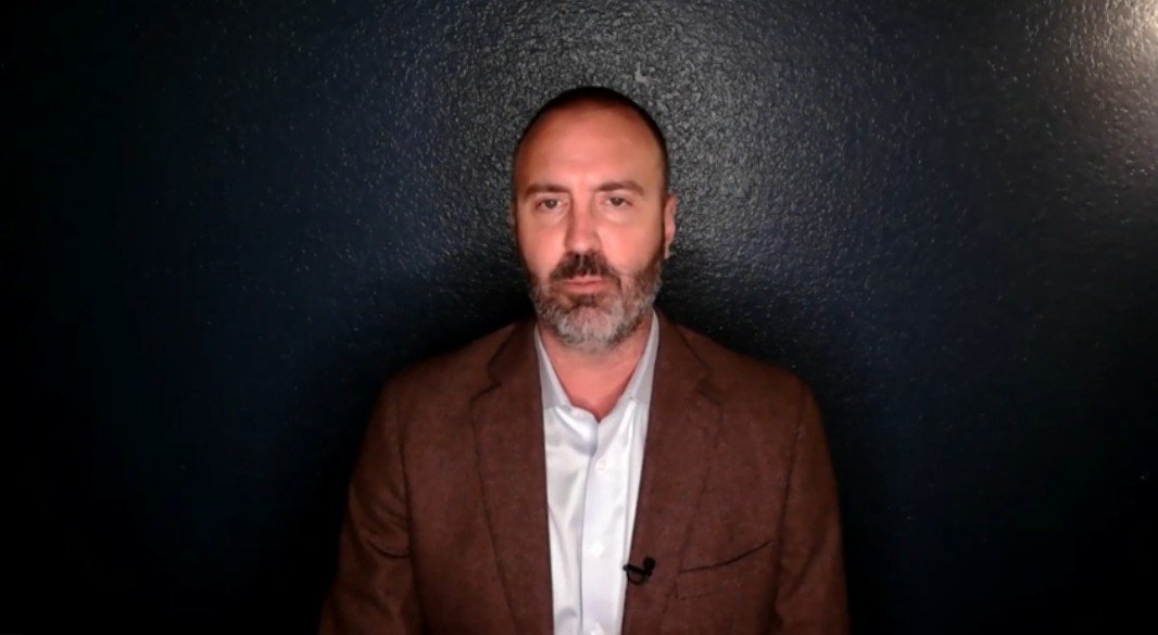 Man with a beard wearing a brown blazer and white shirt, seated against a dark blue wall, presenting a serious expression.