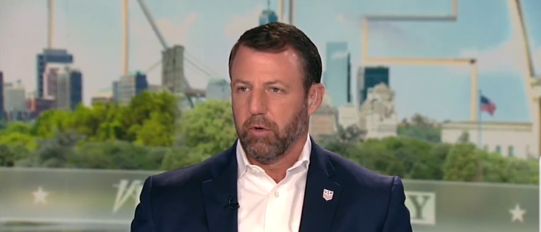 Man in a suit speaking during a television interview with a city skyline in the background, discussing current events and issues.