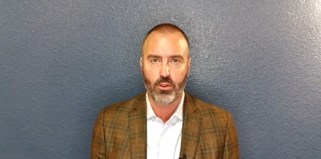 Man in a brown plaid blazer stands against a blue wall, speaking directly to the camera in a professional setting.