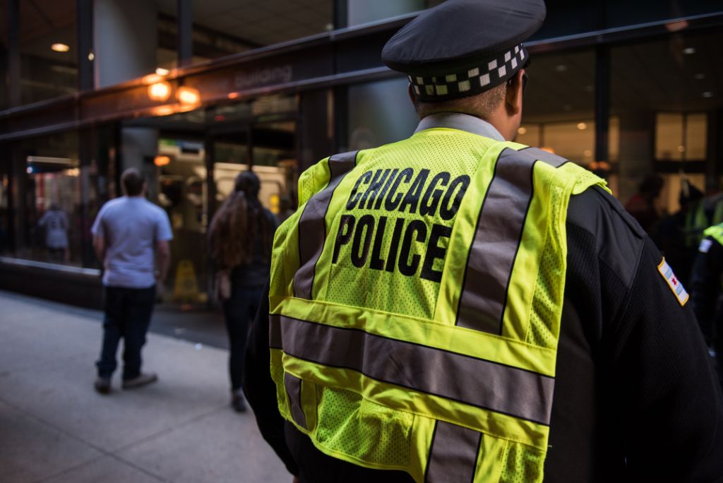 Chicago police officer in a high-visibility vest stands in an urban setting, observing pedestrians in a busy downtown area.