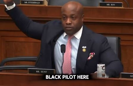 A Black pilot speaking passionately during a congressional hearing, raising his hand to make a point, with a soda can on the table.